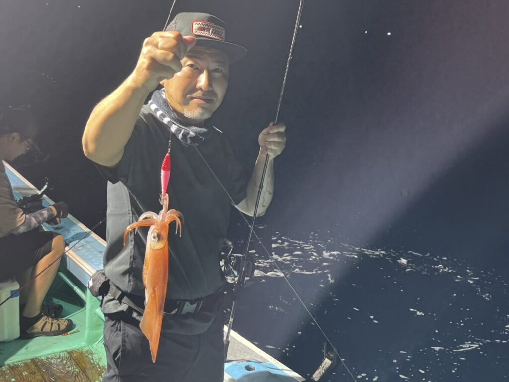 ボートに乗った男がイカのついた釣り糸を垂らし、夜のカメラに向かってポーズをとっている。ボートの背景にはもう一人の人物が座り、その背後には海が見える。
