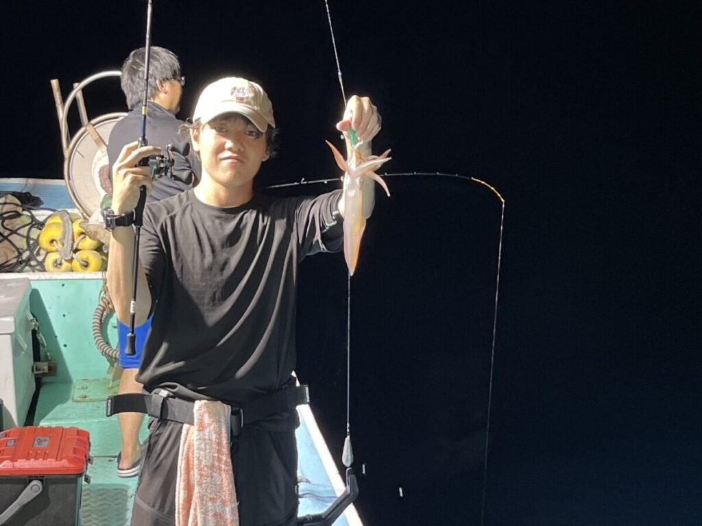 夜の船上で、釣りたてのイカを片手で持ち上げ、もう片方の手には釣り竿を持つ人。背後にはもう一人、向こうを向いて座っている。人工の光に照らされた光景。.