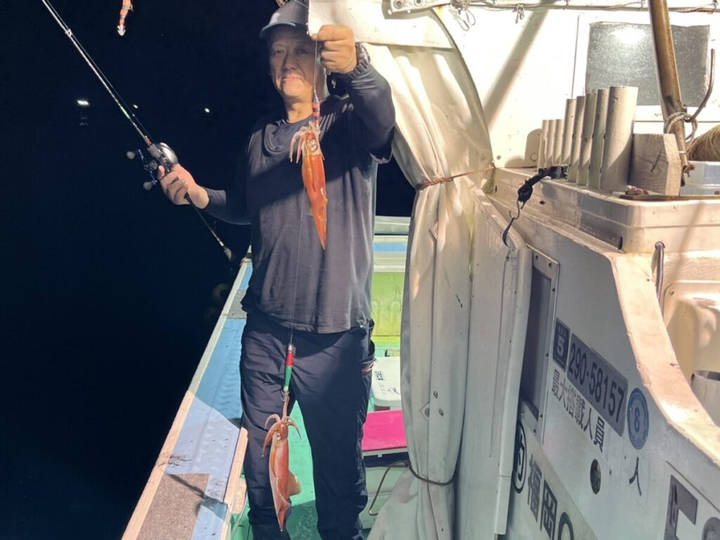 片手に釣り竿を持ち、もう片方の手には釣りたてのイカを2匹掲げている。