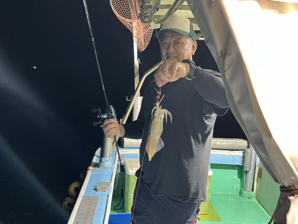 夜のボートの上に立つ男が、明るい照明の下、片手に釣り竿、もう片方の手に釣りたてのイカを掲げ、カメラに向かって微笑んでいる。