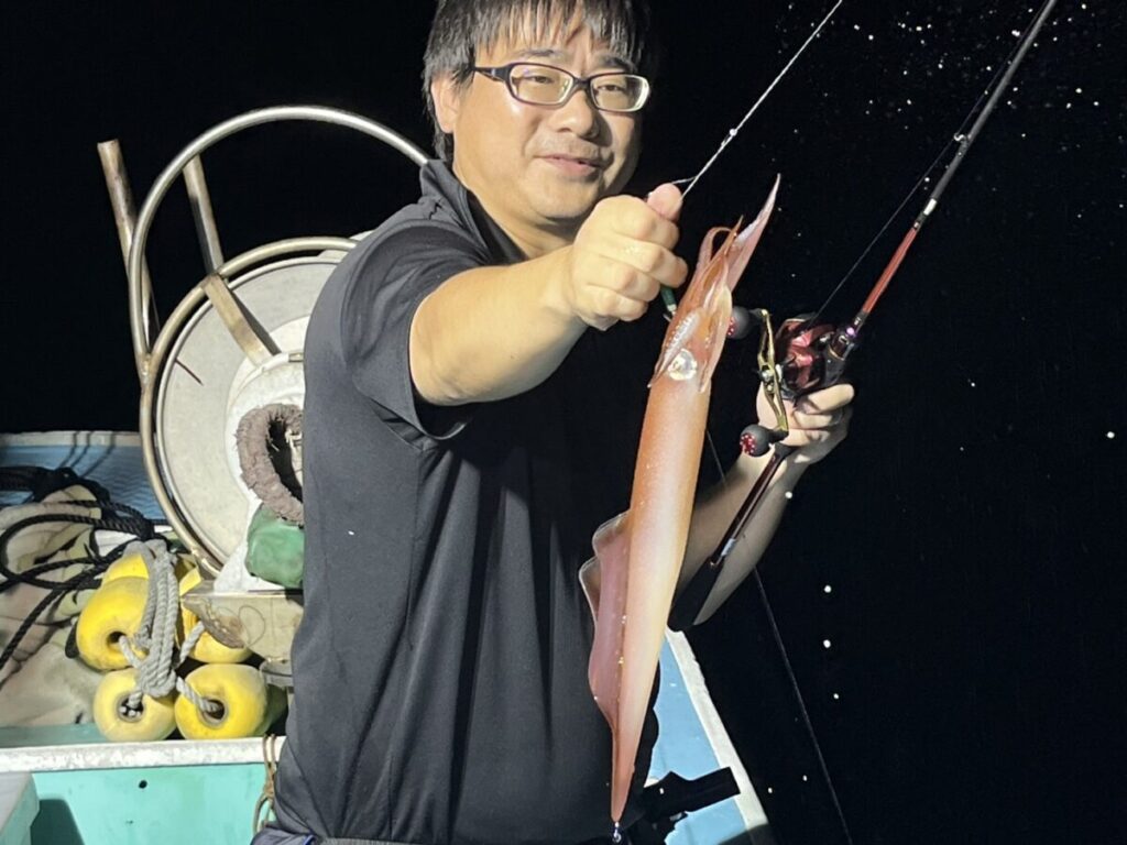 釣り竿を持ち、片手で釣りたてのイカを誇らしげに掲げる夜の船の男。背景には船の設備と暗い海が見える。
