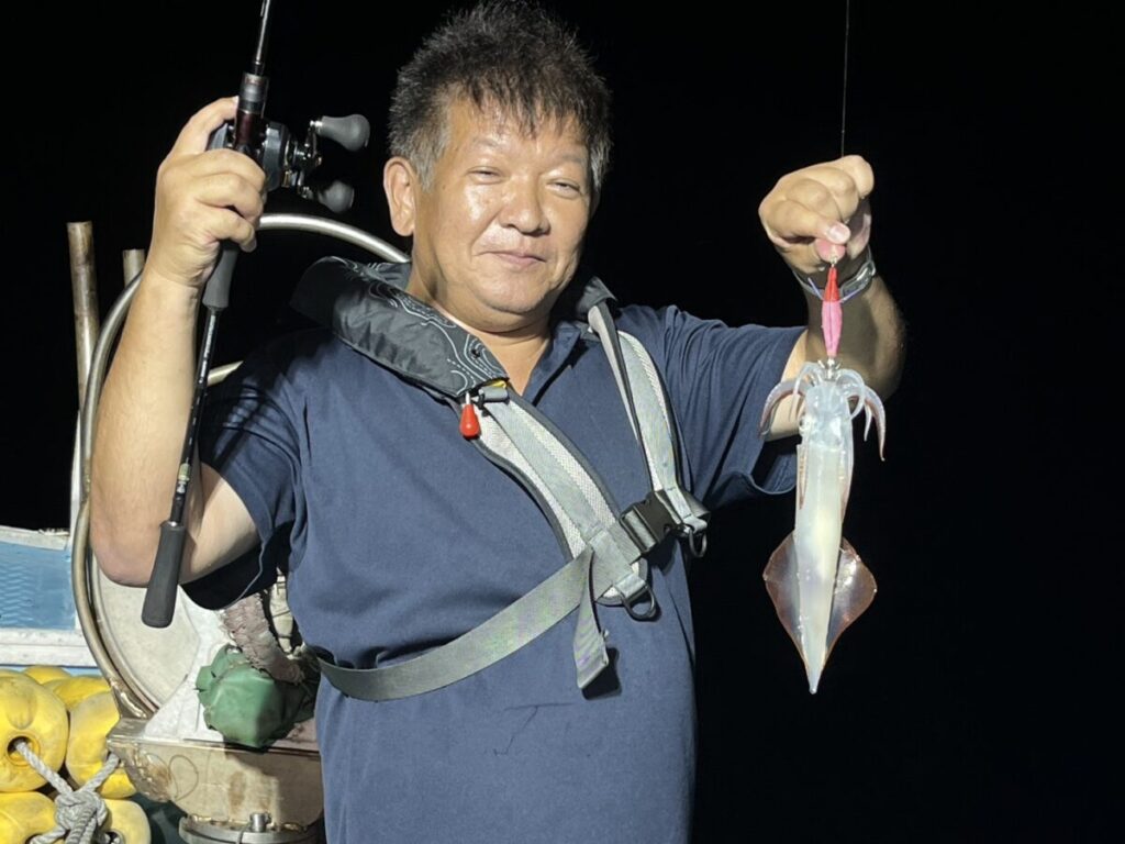 ネイビーのシャツにライフジャケットを着た男が、片手に釣り竿、片手に釣りたてのイカを持ち、夜のボートに立って微笑んでいる。