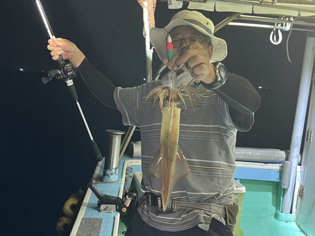 帽子に縞模様のシャツを着た人が、夜の船の上で釣り竿で釣った大きなイカを掲げている。.