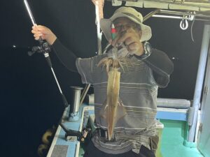 帽子に縞模様のシャツを着た人が、夜の船の上で釣り竿で釣った大きなイカを掲げている。.