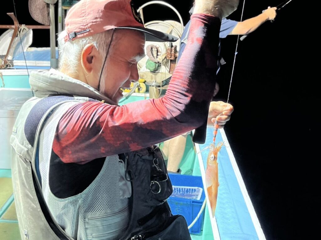 赤い帽子に柄物の長袖シャツ、救命胴衣を着た男が、夜の船上でオレンジ色の小さなイカがかかった釣り糸を持ちながら微笑んでいる。背後では別の人が釣りをしている。