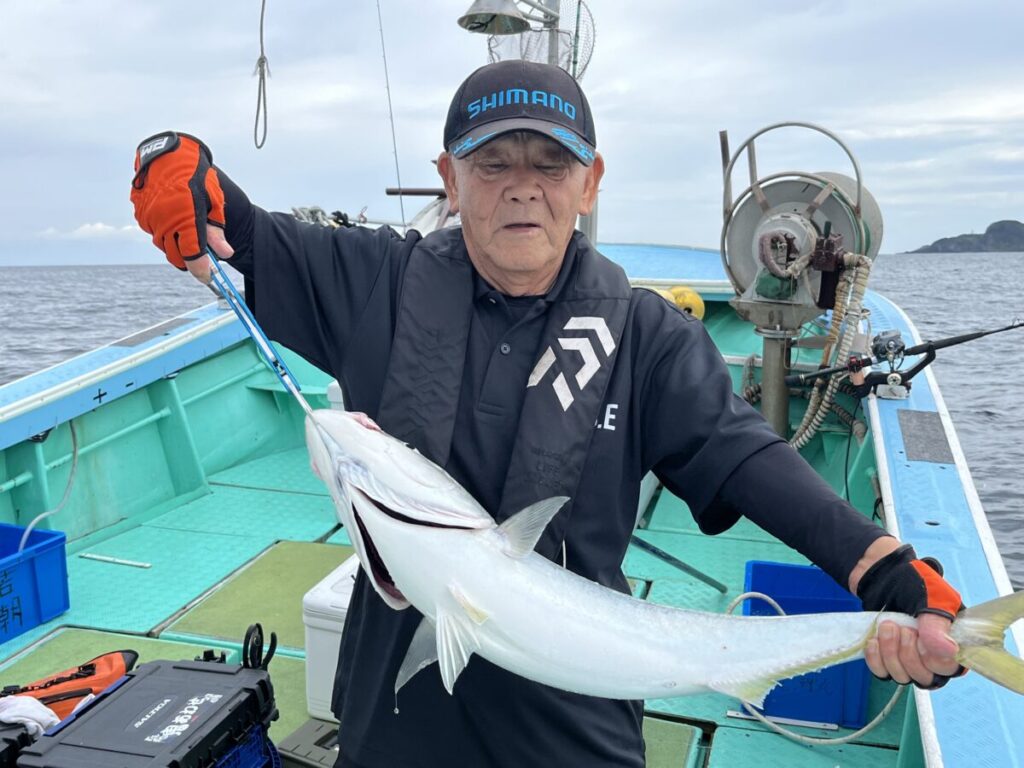 黒い帽子をかぶり、手袋をはめた年配の男性が、ボートの上でフィッシュグリッパーを使って大きな魚を持っている。ボートは水上にあり、漁具が見え、背景には曇り空が広がっている。