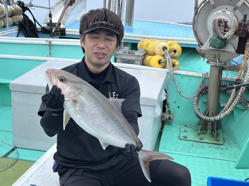 黒い服に手袋をはめた男がボートに乗り、両手で大きな魚を持っている。背景には漁具とロープが見える。
