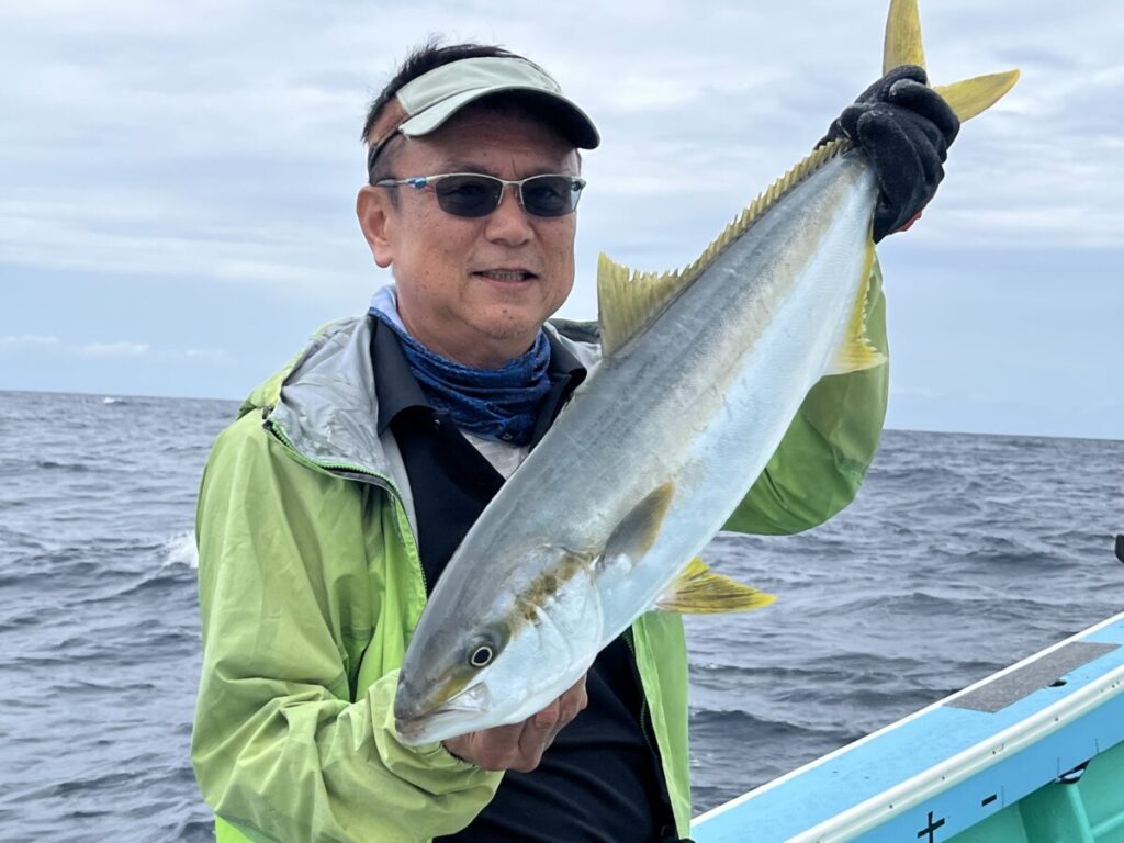 サングラスをかけ、バイザーをかぶり、緑色のジャケットを着た男が、海と曇り空を背景に、ボートの上で大きなブリを手にしている。