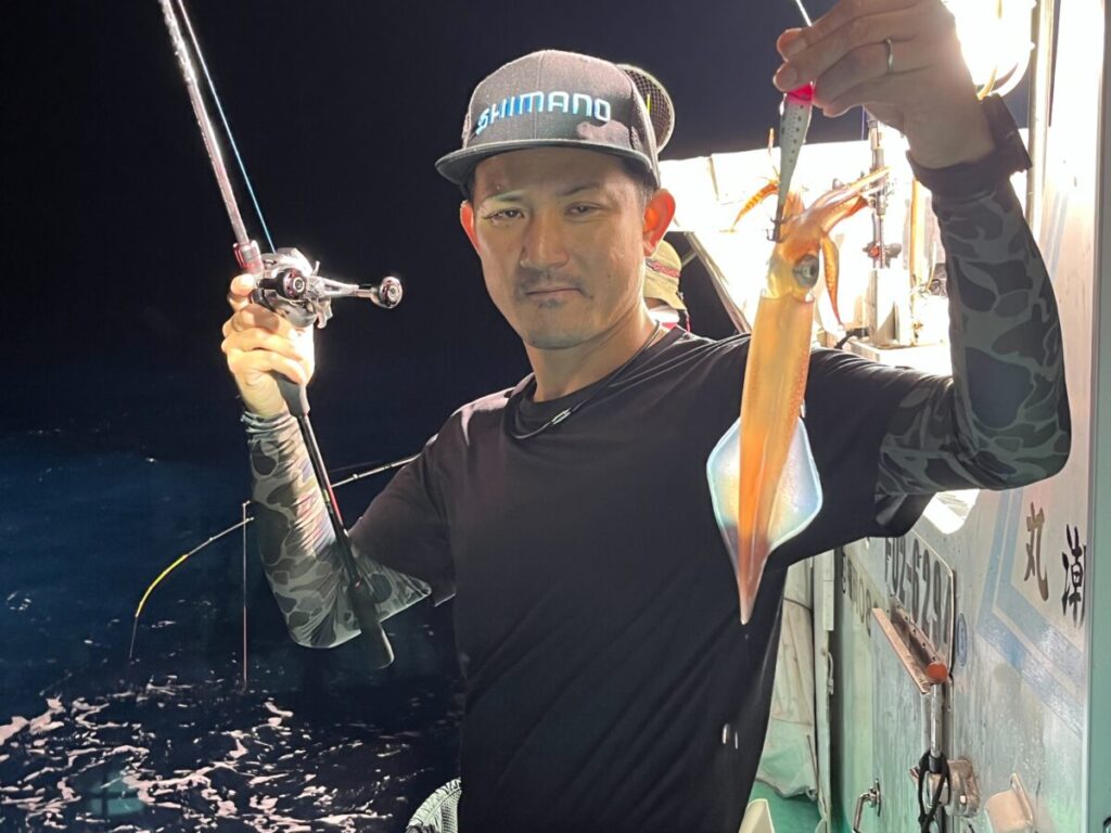 シマノのキャップをかぶり、黒い釣り道具を身につけた男が、片手に釣り竿、もう片方の手には釣りたてのイカを持って、夜の船の上に立っている。.