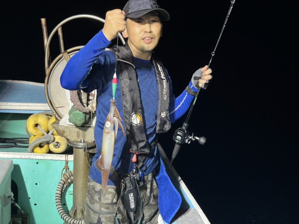 帽子をかぶり、釣り用のベストを着た人が、片手に釣り竿、もう片方の手には釣りたてのイカを持って、夜のボートの上に立っている。背景には船具と暗い夜空が見える。