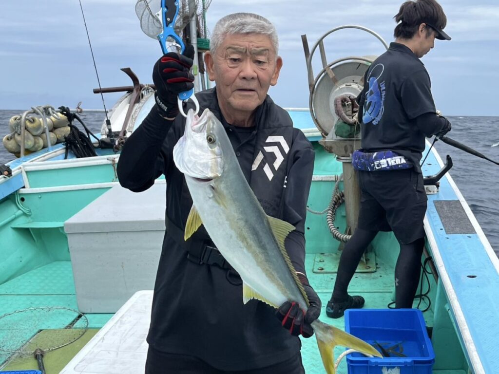 ボートに乗った年配の男性が釣った大きな魚を掲げている。船は曇り空の下、大海原に浮かんでいる。