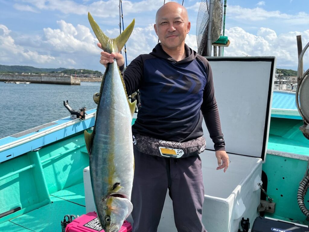 晴れた日、ボートの上で大きな魚を両手で持ち上げる笑顔の男性。背景には大海原と港が見える。ボートの側面はターコイズブルーで、近くには漁具が置かれている。