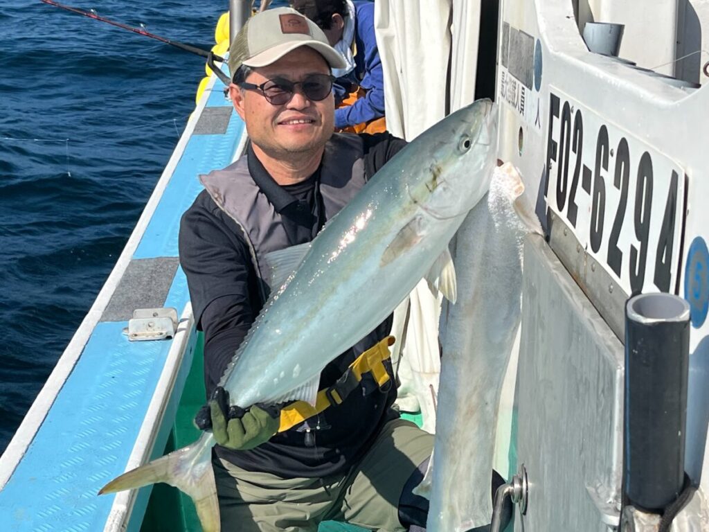 眼鏡をかけ帽子をかぶった人物が、大きな魚を抱いてボートに乗り、カメラに向かって微笑んでいる。