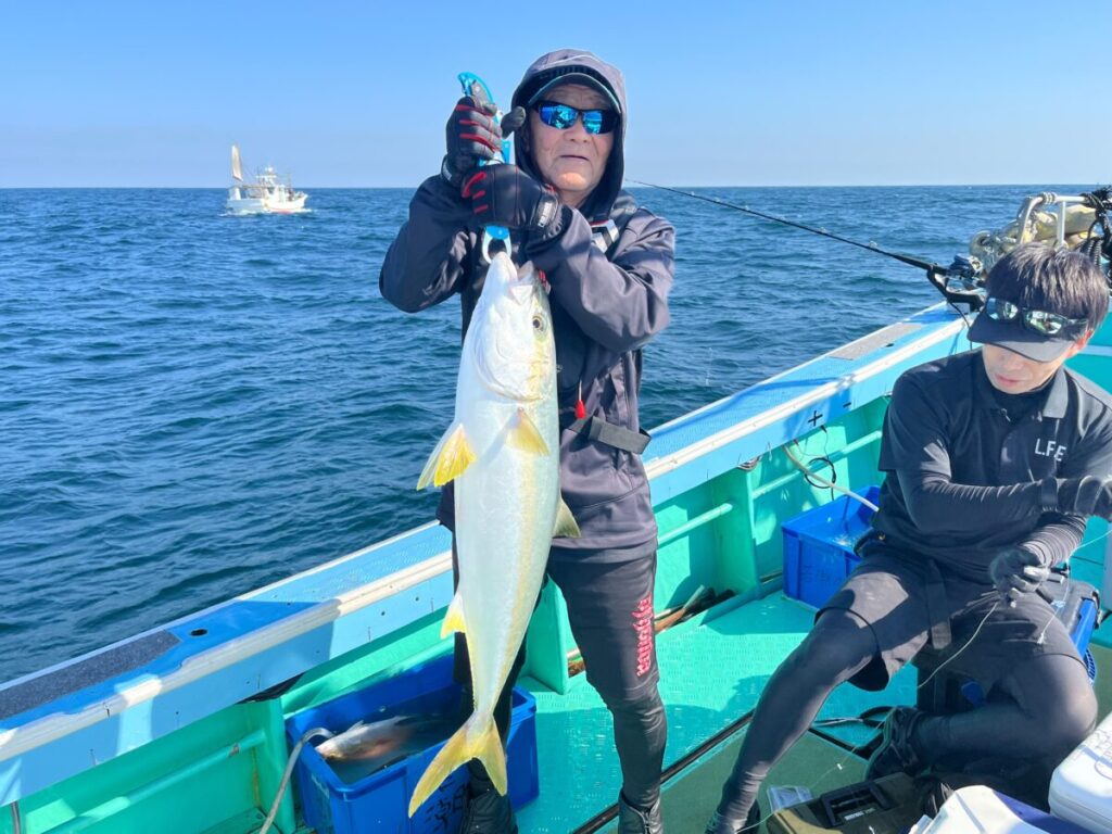 黒っぽい服装にサングラスの人物がボートの上で大きな魚を掲げている。