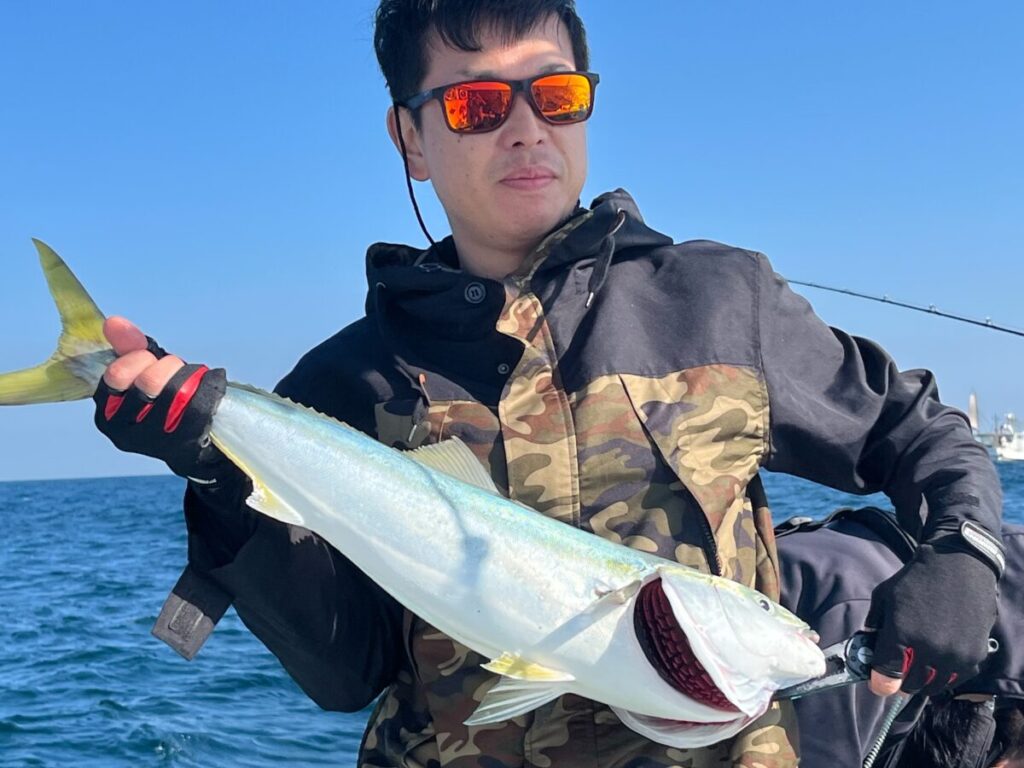 サングラスをかけ、迷彩柄のジャケットを着た人が、黄色い尾を引く大きな魚を手に、澄み切った青空の下、海に浮かぶボートの上に立っている。
