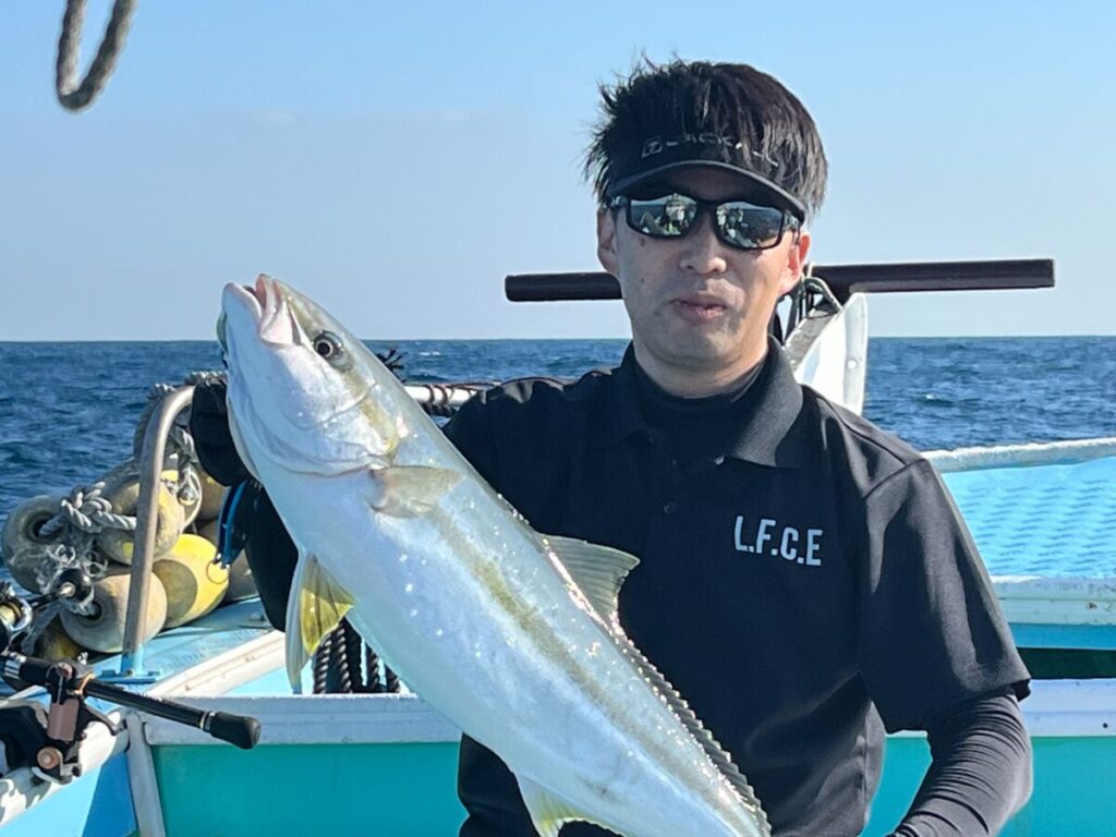 サングラスをかけ、黒いシャツを着た人物が、海と青空を背景に、ボートの上で大きな魚を持っている。