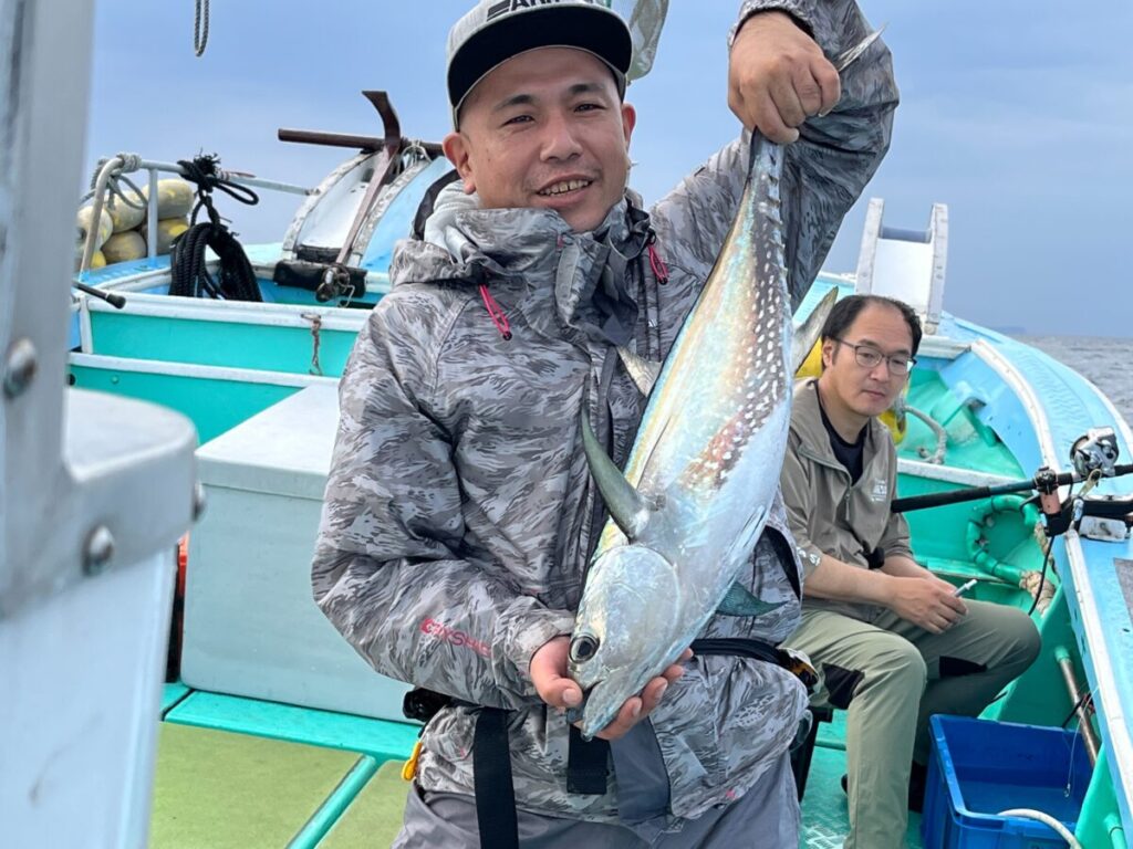 グレーのジャケットに帽子をかぶった笑顔の男性が、漁船で大きな魚を手にしている。