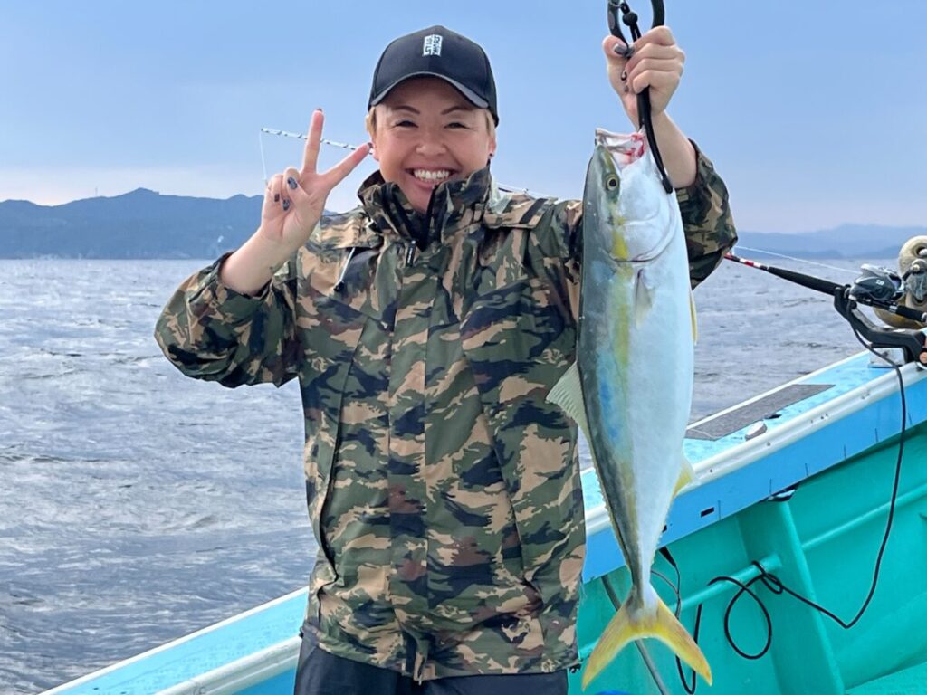 迷彩柄のジャケットに黒い帽子の笑顔の人物が、ボートの上で大きな魚を掲げ、片手でピースサインをしている。.