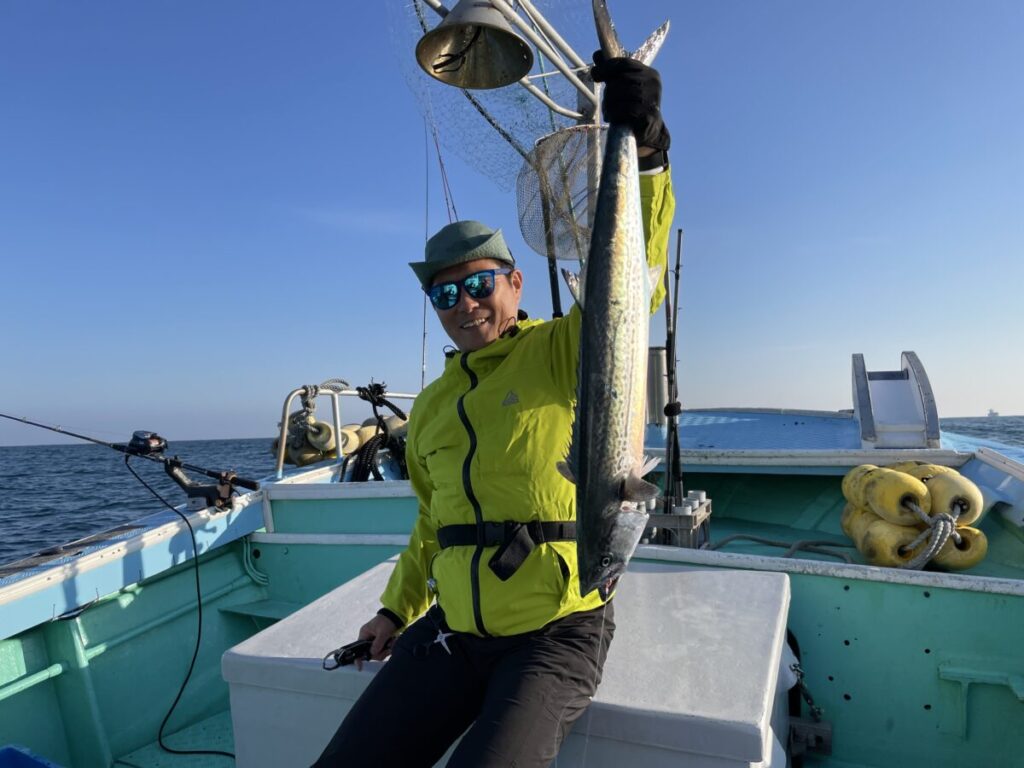 サングラスをかけ、緑色のジャケットを着た人物が、漁具と青空を背景に、海に浮かぶボートの上で大きな魚を掲げて微笑んでいる。.