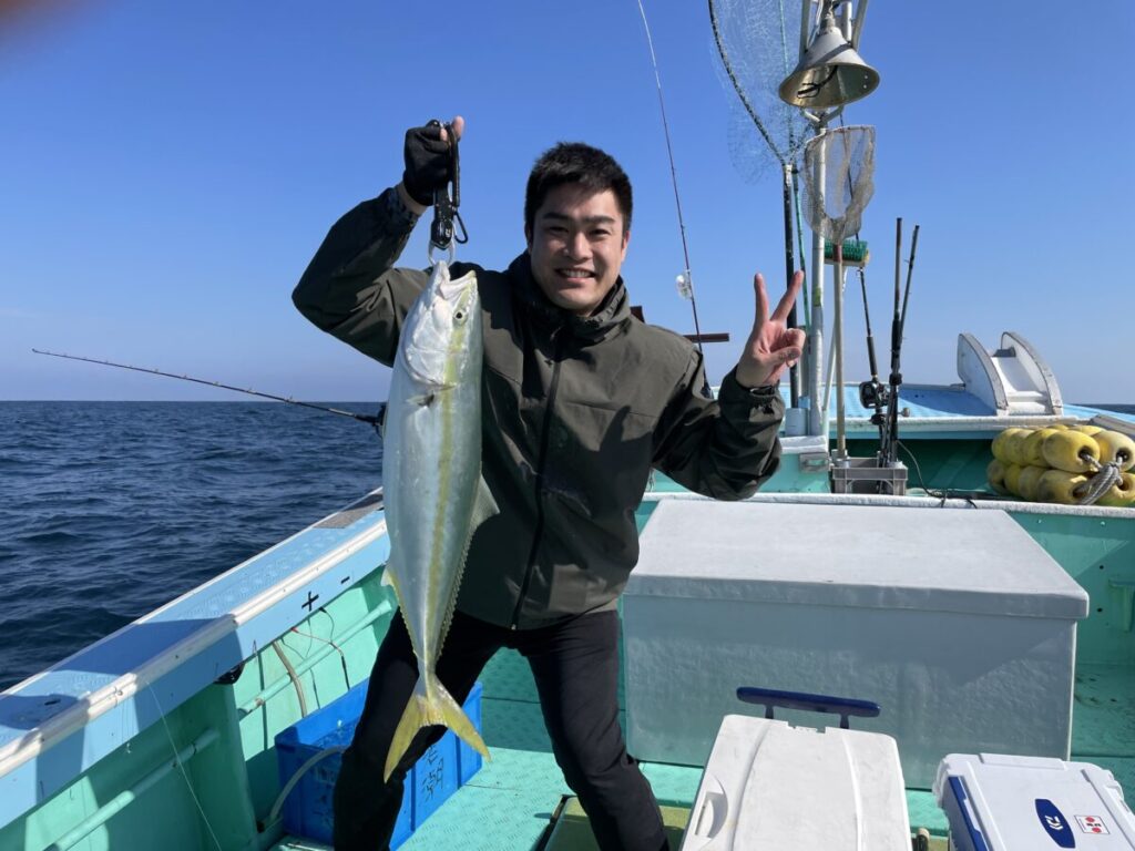 ボートの上で、片手で大きな魚を掲げ、もう片方の手でピースサインをする笑顔の人。背景には海と青い空、そして船の上の漁具が見える。.