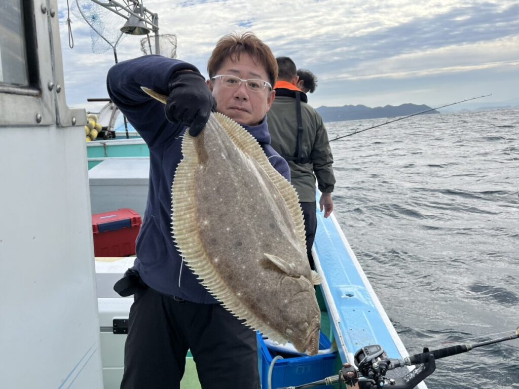 曇り空の下、ボートに乗った人が大きなヒラメと思われる魚を掲げている。.