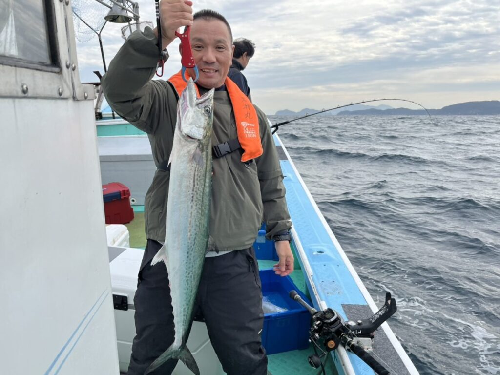 ボートの上で大きな魚を片手で持ち上げる笑顔の男性。緑のジャケットにオレンジの救命胴衣。曇り空の下、彼の周りには漁具と荒波が見える。.