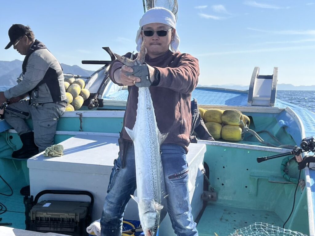 サングラスをかけ帽子をかぶった人物が、ターコイズブルーの漁船の上で大きな魚を掲げている。空は晴れ渡り、海は穏やかだ。.