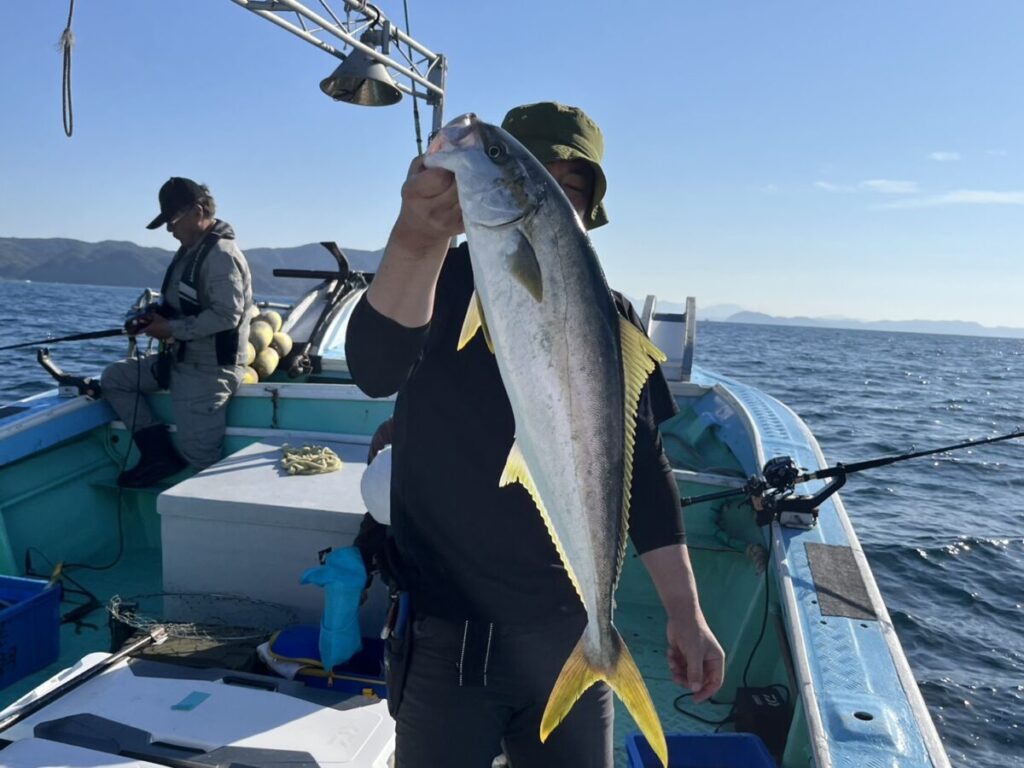 ボートに乗った人が尾の黄色い大きな魚を掲げ、後ろで別の人が漁具の準備をしている。.