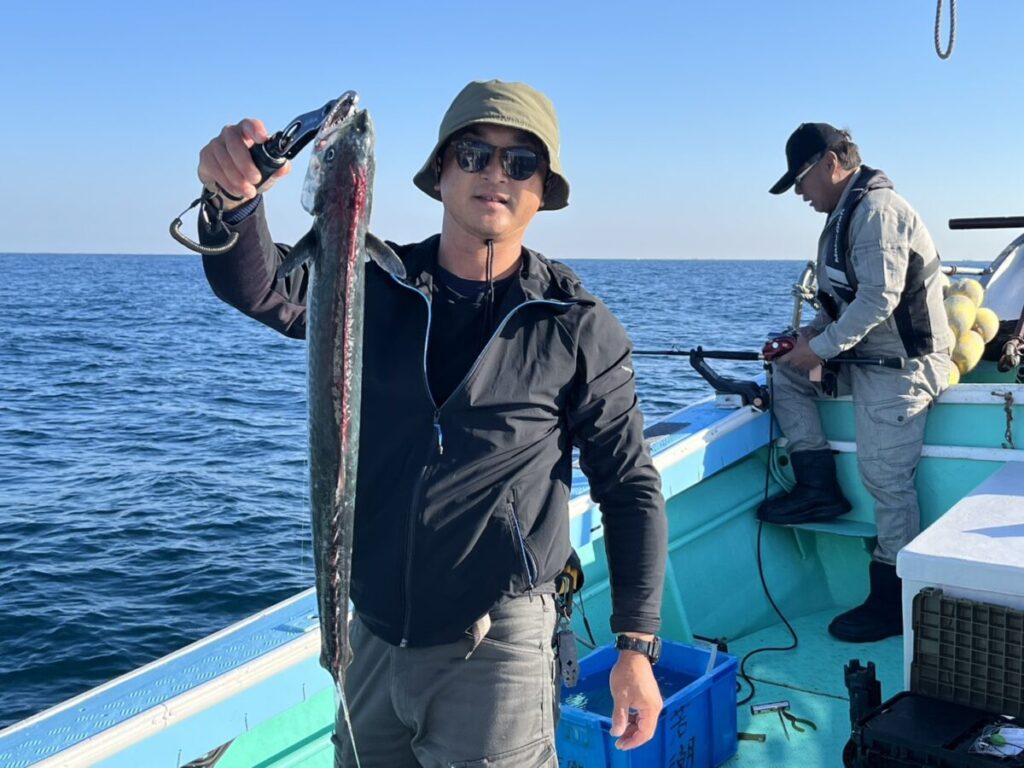 サングラスをかけ、帽子をかぶった人物がボートの上で大きな魚を掲げている。.