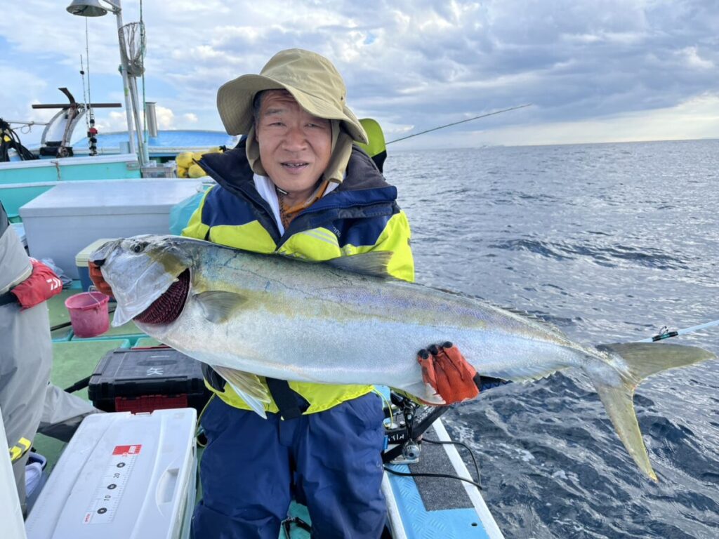 海と曇り空を背景に、黄色と青のジャケットに帽子、手袋をはめた人物がボートに座り、大きな魚を抱いて微笑んでいる。.