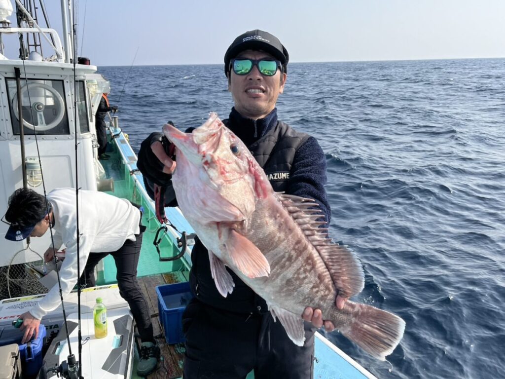 サングラスをかけ、帽子をかぶった人物が、海上で大きな獲れたての魚を誇らしげに手にしている。海と澄んだ空が見える。.