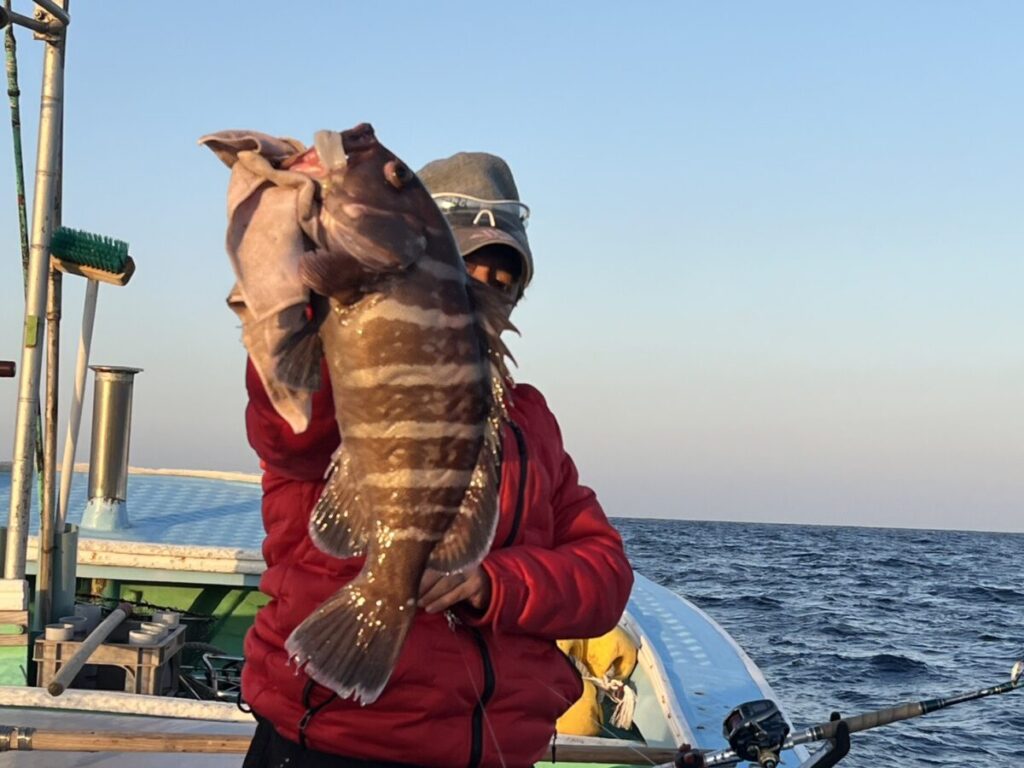 海と澄み切った空を背景に、赤いジャケットと帽子を被った人物が、ボートの上で大きな縞模様の魚を掲げている。.