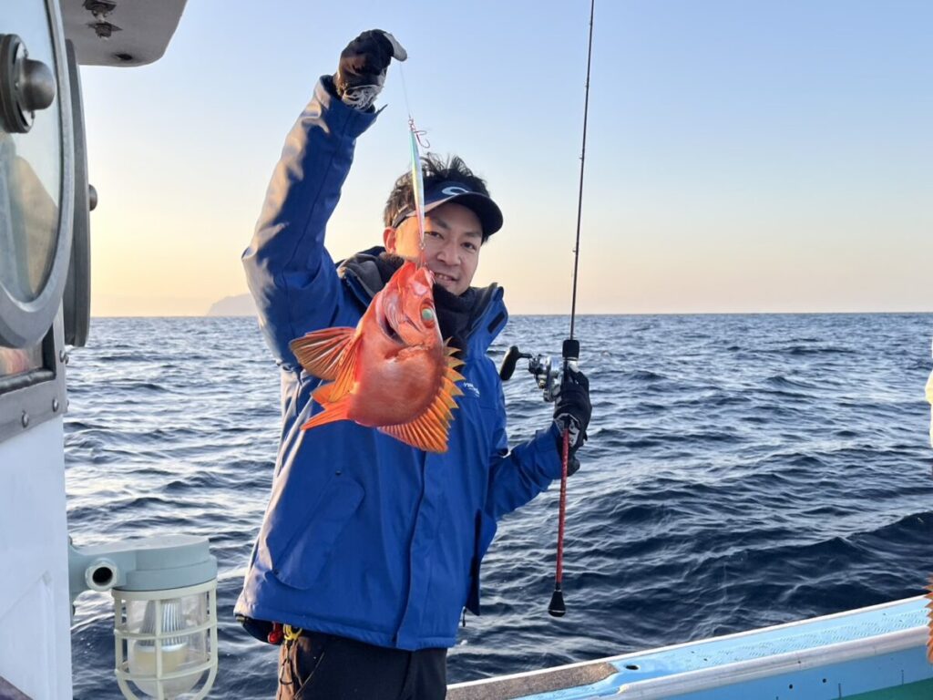 海と澄み切った空を背景にしたボートの上で、鮮やかなオレンジ色の魚を釣り糸に掲げながら微笑む青いジャケットと帽子の人物。.