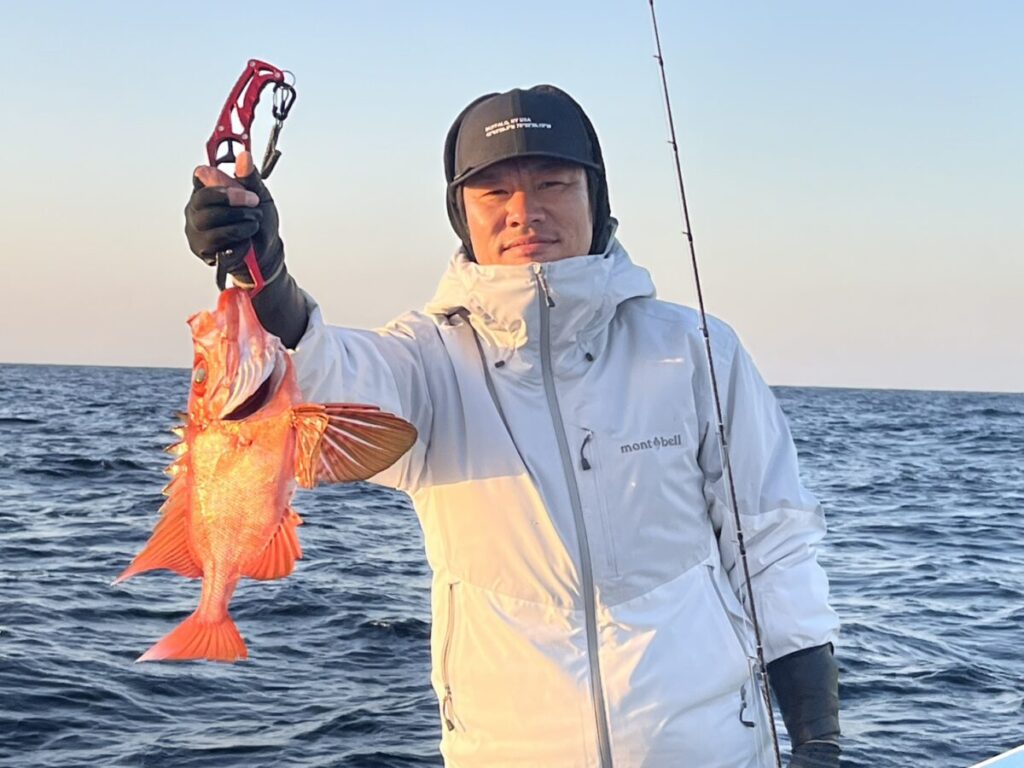 白い上着に黒い帽子の人物が、オレンジ色でとがったヒレの大きな魚を掲げ、釣り竿を持ったボートの上に立っている。.