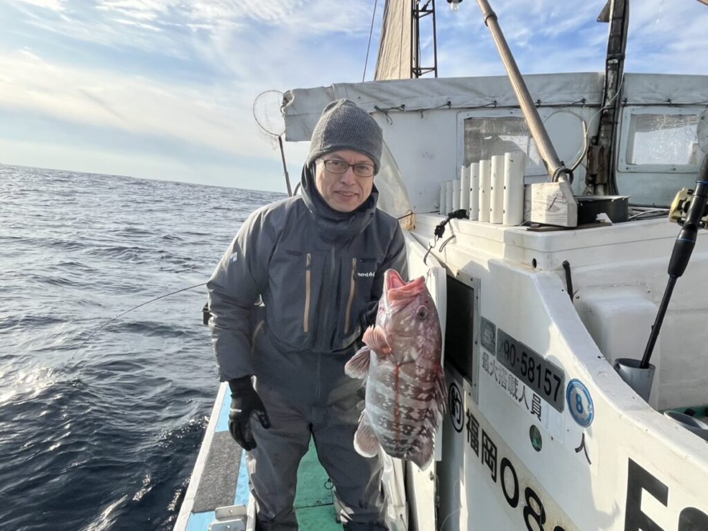 眼鏡をかけ、防寒着を着た人物が、海と空を背景に、釣りたての大きな魚を手にボートの上に立っている。.