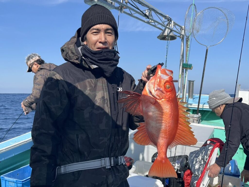 黒っぽいジャケットにビーニーをかぶった男性が、ボートの上で鮮やかなオレンジ色の大きな魚を持っている。澄み切った青空の下、背景では他の2人が釣りをしている。ボートの上には漁網と漁具が見える。.