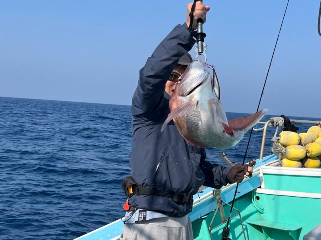 晴れた日の海と漁具を背景に、ボートに乗った人が大きな魚を釣り針で支えている。.