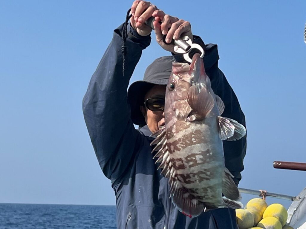 海と澄み切った青空を背景に、サングラスをかけ、フード付きのジャケットを着た人物が、ボートの上で大きな柄の魚をペンチで掲げている。.