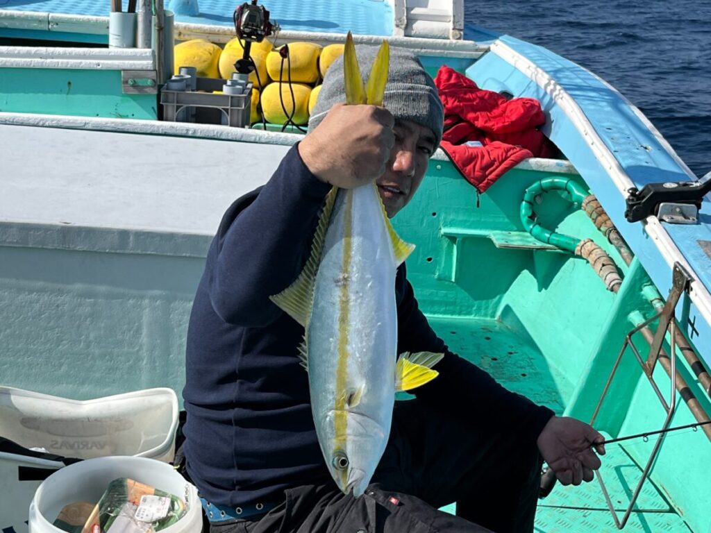 ターコイズブルーのボートに腰掛けた人が、片方の手で大きなブリを掲げている。船上には漁具と黄色い浮き輪が見え、背景には海が広がっている。.
