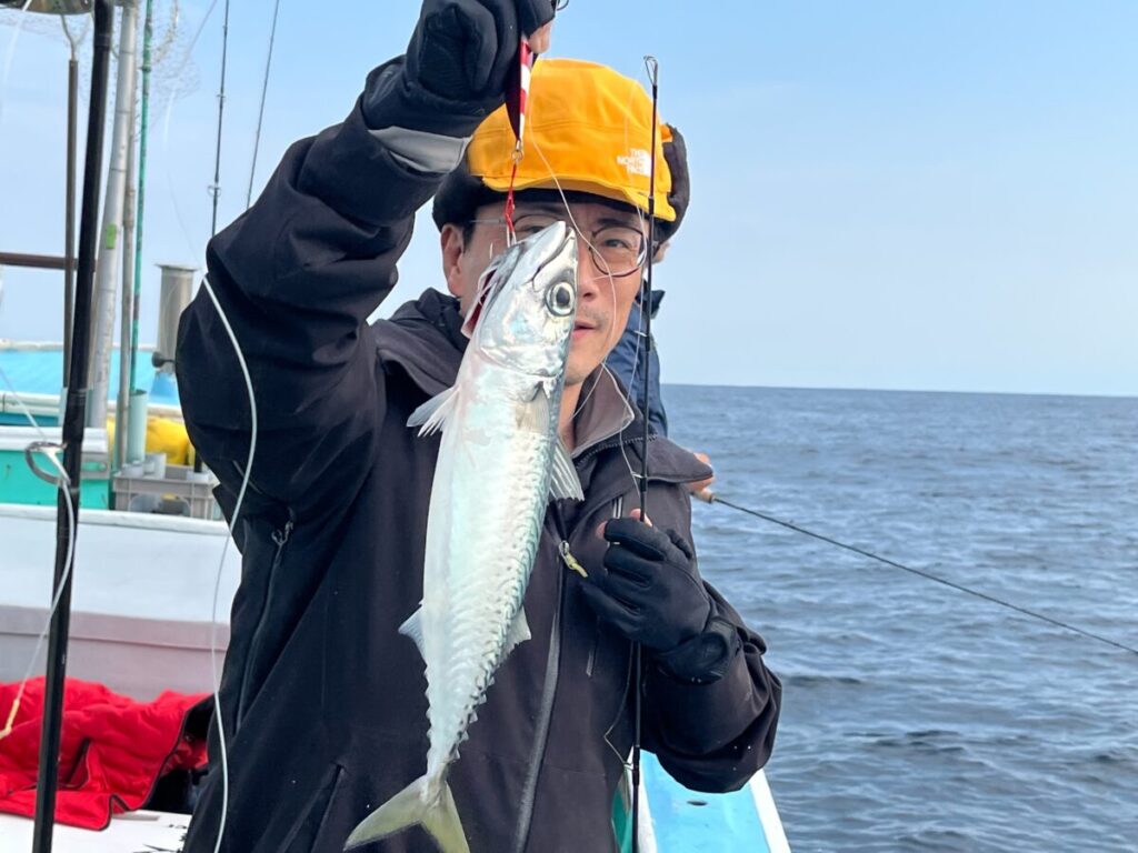 黄色いヘルメットをかぶり、黒い手袋をはめた人が船の上で釣りたての魚を掲げている。.