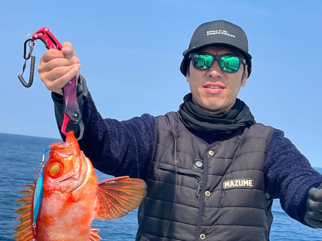 サングラスをかけ、黒い帽子をかぶり、黒いベストを着た人物が、海と青空を背景にボートの上に立ちながら、大きな目をした鮮やかなオレンジ色の魚を掲げている。.