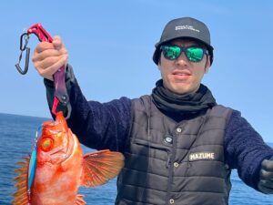 サングラスをかけ、黒い帽子をかぶり、黒いベストを着た人物が、海と青空を背景にボートの上に立ちながら、大きな目をした鮮やかなオレンジ色の魚を掲げている。.