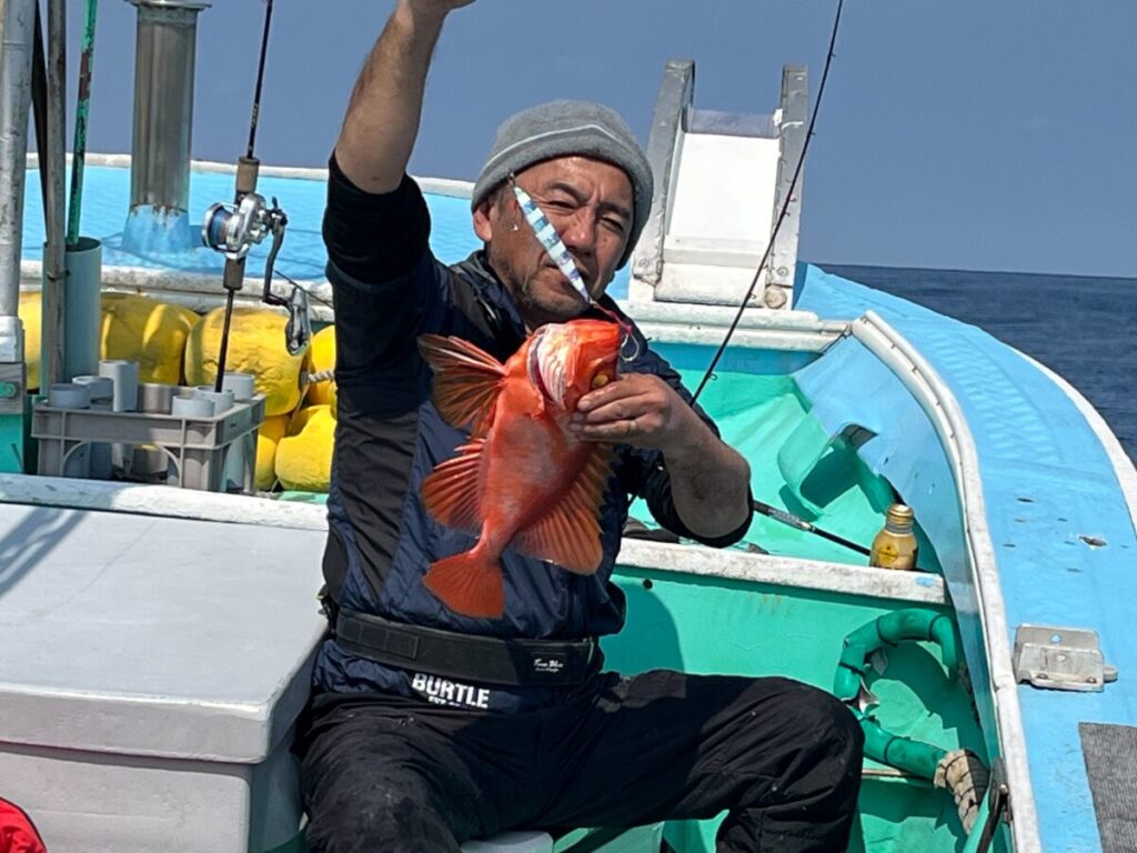 小舟に腰掛けた男が、釣ったばかりの鮮やかなオレンジ色の魚を掲げ、誇らしげにカメラに向かって微笑んでいる。背景には海と空が見える。.