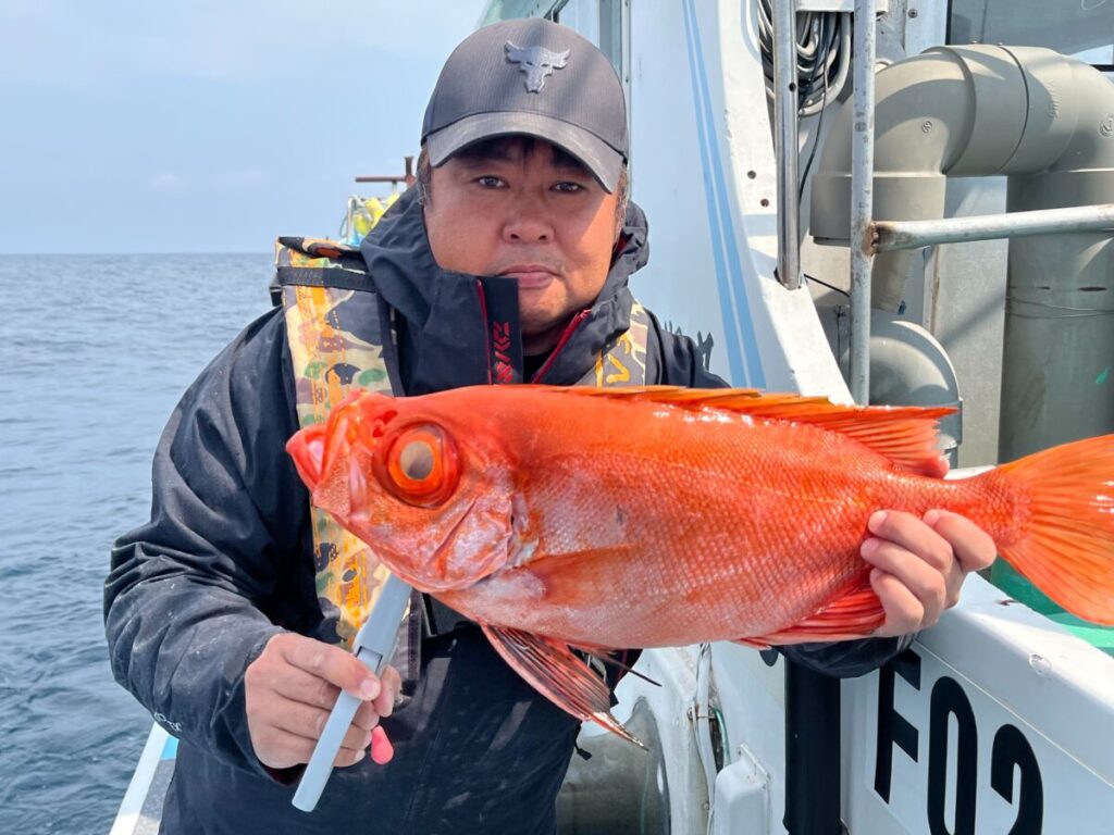 ボートの上に立つ人が、目の目立つ鮮やかなオレンジ色の大きな魚を抱いている。背景には海と空が見える。その人物は黒い帽子をかぶり、救命胴衣のついたジャケットを着ている。.