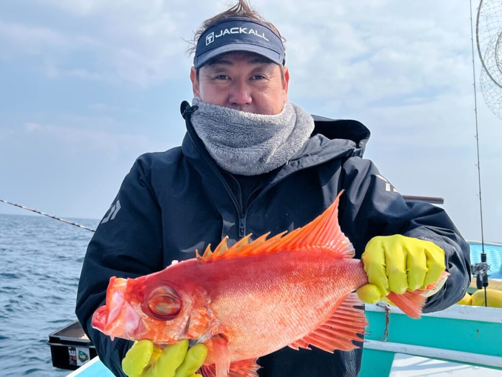 海と空を背景にしたボートの上で、黒いジャケットに黄色の手袋をはめた人物が真っ赤な大きな魚を持っている。その人物はスカーフを巻き、バイザーの帽子をかぶっている。.