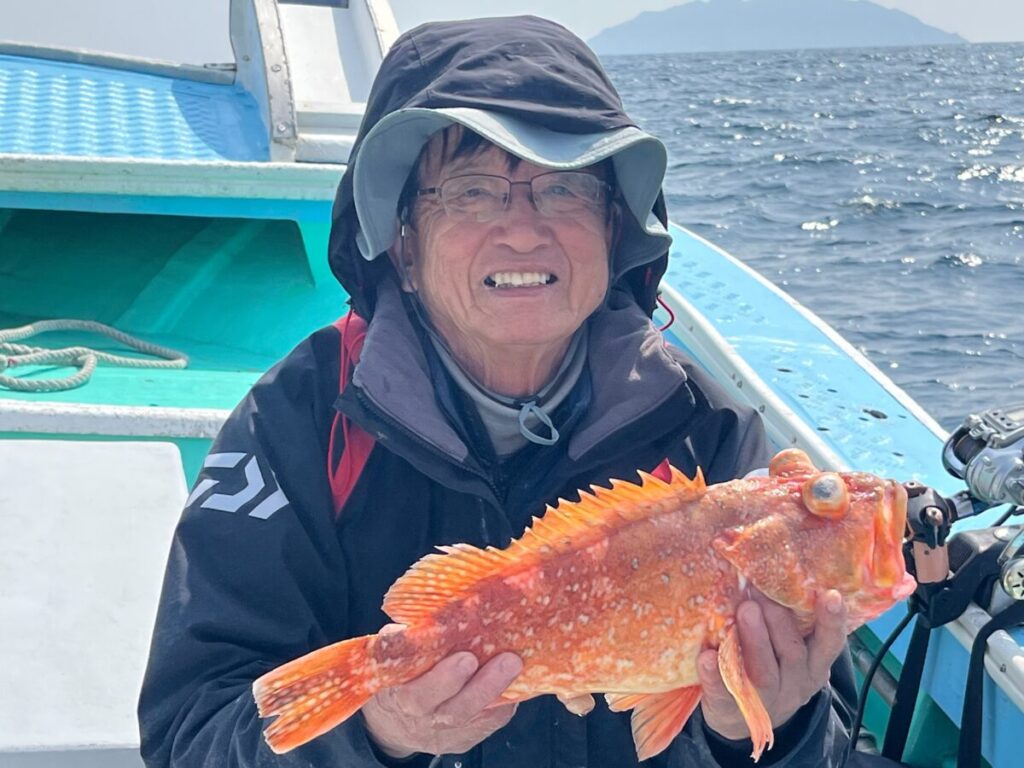 眼鏡をかけ、フード付きのジャケットを羽織った年配の人物が、水と遠くの島を背景に、海上のボートの上でオレンジ色の大きな魚を手にして微笑んでいる。.