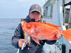 黒いジャケットに帽子をかぶった人物が、大きな目をした鮮やかなオレンジ色の大きな魚を抱いてボートの上に立っている。背景には海とボートの一部が見える。.