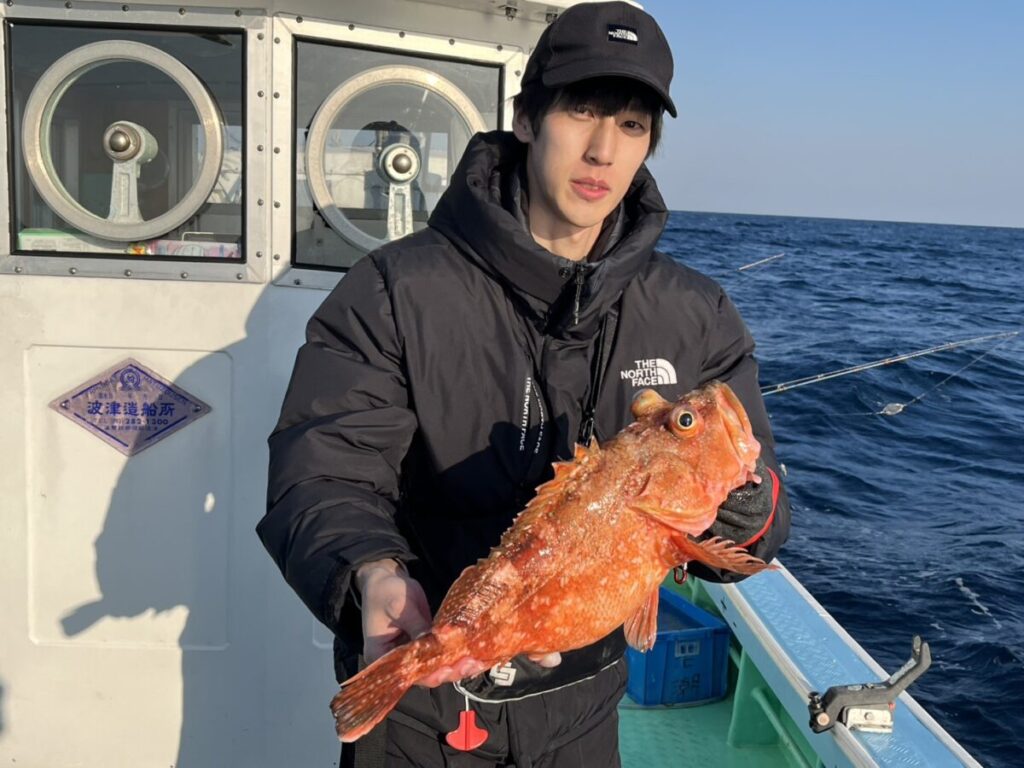 釣り竿を背景に、頭上には青空が広がる海上で、黒いジャケットと帽子を被った人物がオレンジがかった赤色の大きな魚を手にしている。.