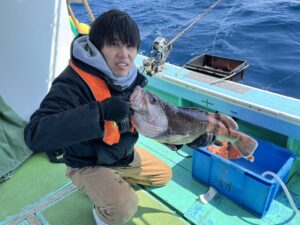 ボートに乗った人が両手で大きな魚を持ち、カメラに向かって微笑んでいる。海を背景に釣り竿と青い容器が見える。.