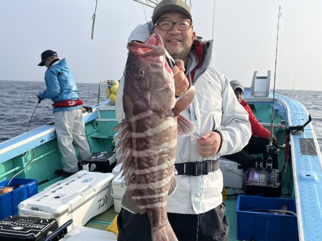 眼鏡をかけ帽子をかぶった笑顔の人物が、船上で大きな縞模様の魚を誇らしげに手にしている。.